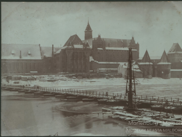 Fotografia przedstawia zimowy obraz zachodniej części zespołu zamku krzyżackiego. Na pierwszym planie znajduje się zamarznięta rzeka Nogat z kładką pieszą łączącą dwa brzegi. W tle zaśnieżone dachy zamkowe.
