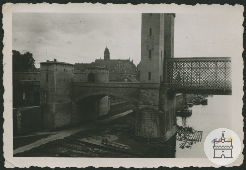 Fotografia przedstawia bramę mostową – wjazd na most drogowy na Nogacie po północnej stronie zamku krzyżackiego. W tle widoczne są zabudowania zamkowe.