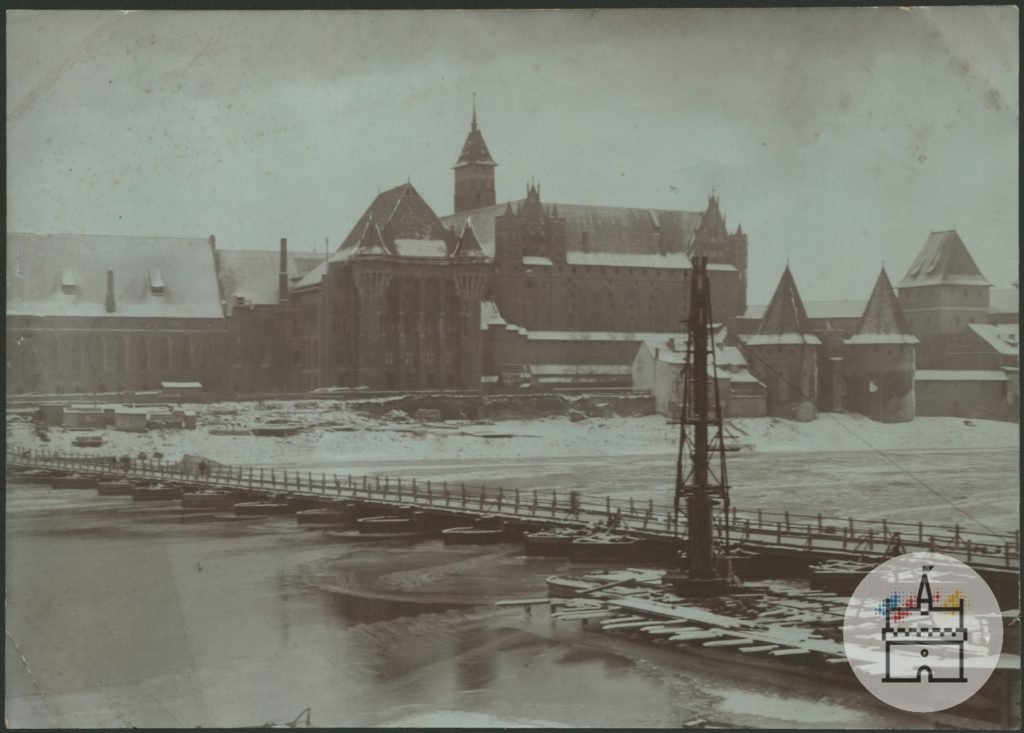 Fotografia przedstawia zimowy obraz zachodniej części zespołu zamku krzyżackiego. Na pierwszym planie znajduje się zamarznięta rzeka Nogat z kładką pieszą łączącą dwa brzegi. W tle zaśnieżone dachy zamkowe.